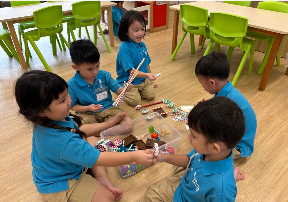 Five children in blue uniforms sit on a classroom floor, sharing candy-shaped toys from a clear bin.